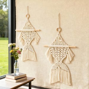 May include: Two off-white macrame wall hangings. Each features a triangular top, a wooden dowel, and a fringed bottom. The hangings are suspended by a wooden ring and a length of cord. The background is a textured, off-white wall.