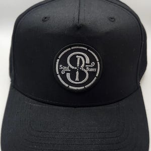 May include: Black baseball cap with a curved brim. A circular patch on the front displays the words "Soul Bones" around a stylized "SB" design in white. The cap is made of a textured fabric.