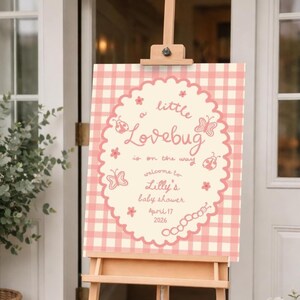 May include: A baby shower sign on a wooden easel. The sign has a pink and white gingham background with a scalloped border. Text reads "a little Lovebug is on the way" and welcomes guests to Lilly's baby shower, April 17, 2026.