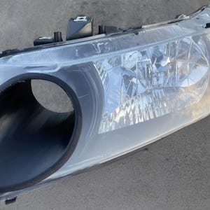 May include: A clear automotive headlight assembly with a black housing. The headlight features a large, round opening and a clear lens. The assembly is designed for a vehicle and is likely a replacement part.