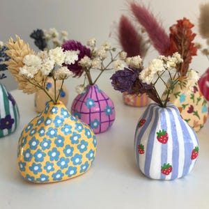 Può includere: Una collezione di piccoli vasi in ceramica dipinti a mano, ognuno decorato in modo unico con motivi floreali e a righe in vari colori. I vasi contengono fiori secchi, creando un'affascinante esposizione decorativa.
