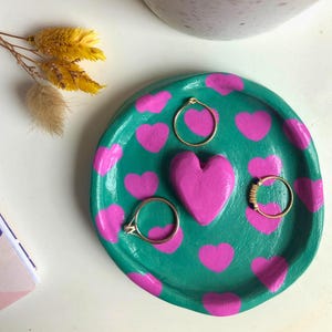 Könnte beinhalten: Eine türkisfarbene Schmuckschale mit rosa Herzmustern und einem großen rosa Herz in der Mitte. Drei goldene Ringe sind auf der Schale ausgestellt. Ein weiß-rosa gesprenkelter Topf befindet sich im Hintergrund.