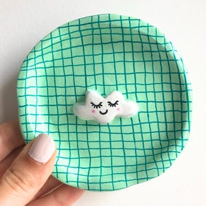 Pode incluir: Um pequeno prato redondo pintado à mão com um padrão de grade verde menta. Uma nuvem branca com olhos fechados e bochechas rosadas está centrada no prato. O prato é segurado por uma pessoa.