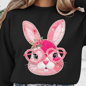 May include: Black sweatshirt featuring a pink bunny face with heart-shaped glasses. The bunny has patchwork details, floral accents, and a pink nose. The design is centered on the front of the black sweatshirt.