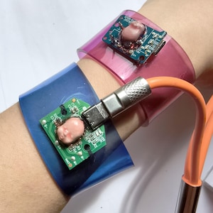 May include: Two translucent plastic cuffs in pink and blue, each featuring a small green circuit board with a miniature doll face attached. An orange charging cable with a silver metal connector is plugged into the blue cuff's circuit board.