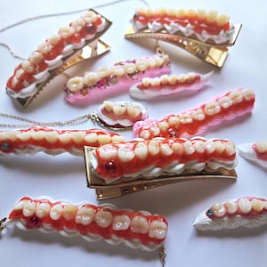 Può includere: Una collezione di fermagli per capelli e gioielli con denti umani in miniatura realistici incastonati in un materiale simile alla panna montata bianca con accenti rossi lucidi e piccoli strass colorati su basi in metallo dorato.