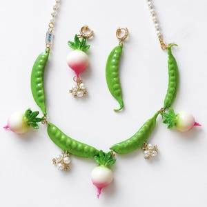 Puede incluir: Un conjunto de joyería que incluye un collar y pendientes con vainas de guisantes verdes, rábanos blancos y rosas, y pequeños racimos de perlas blancas. La cadena del collar es de perlas blancas y los pendientes son aros dorados con cristales.