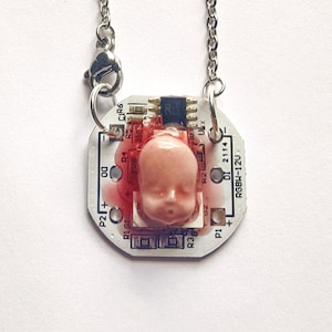 May include: A necklace featuring a white circular circuit board pendant with the text RGBW-12V and DI 2114 printed on it. A small, pale pink, realistic doll head is mounted in the center, surrounded by a translucent red resin-like material.
