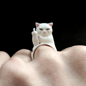 May include: A white ring featuring a sculpted cat figure with pink-tinted ears and nose. The cat is depicted with its eyes closed and one paw raised, extending its middle finger in a gesture. The ring band is white and encircles the finger.