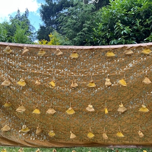 Puede incluir: Un textil marrón dorado con intrincados bordados florales y adornos colgantes en forma de campana. La tela está drapeada, con un fondo natural de follaje verde y un cielo parcialmente nublado.