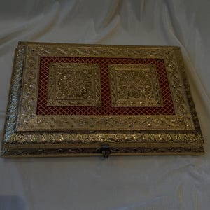 May include: A rectangular, gold-coloured box with intricate embossed detailing. The box features a red, patterned interior with two square, floral-designed panels. The box is closed with a small clasp.