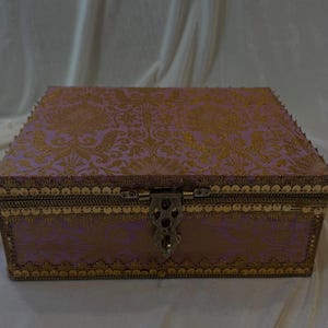 Puede incluir: Una caja rectangular con tapa abisagrada, con una tela lavanda con un estampado floral dorado. La caja está adornada con metal dorado y un cierre decorativo. El diseño general sugiere un estilo vintage o antiguo.