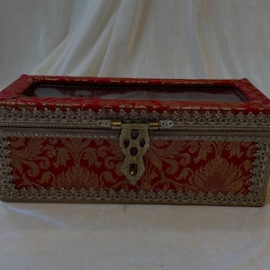 Puede incluir: Caja decorativa rectangular roja y dorada con tapa transparente. La caja presenta un estampado floral y un ribete dorado ornamentado. Un cierre dorado asegura la parte delantera. La caja probablemente se utiliza para guardar o exhibir.