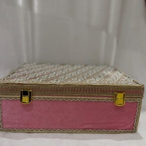 Puede incluir: Una caja de terciopelo rosa con ribete dorado y una parte superior decorativa con cuentas. La caja tiene un cierre dorado y probablemente se utiliza para guardar o regalar. La caja es de forma rectangular.