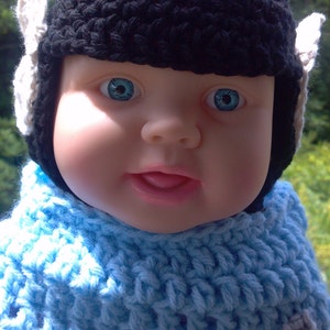 Op de afbeelding: Een babypop met een zwarte gehaakte muts met witte oorflappen en een lichtblauwe gehaakte trui.