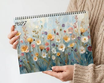 May include: A spiral-bound wall calendar for the year 2027 featuring an impressionistic painting of a wildflower meadow. The artwork displays a variety of yellow, white, pink, and purple flowers against a soft blue and cream background.