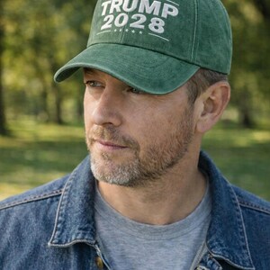May include: Green baseball cap with the text "TRUMP 2028" in white. The cap is worn by a person wearing a denim jacket and a gray t-shirt. The background is a blurred outdoor scene.