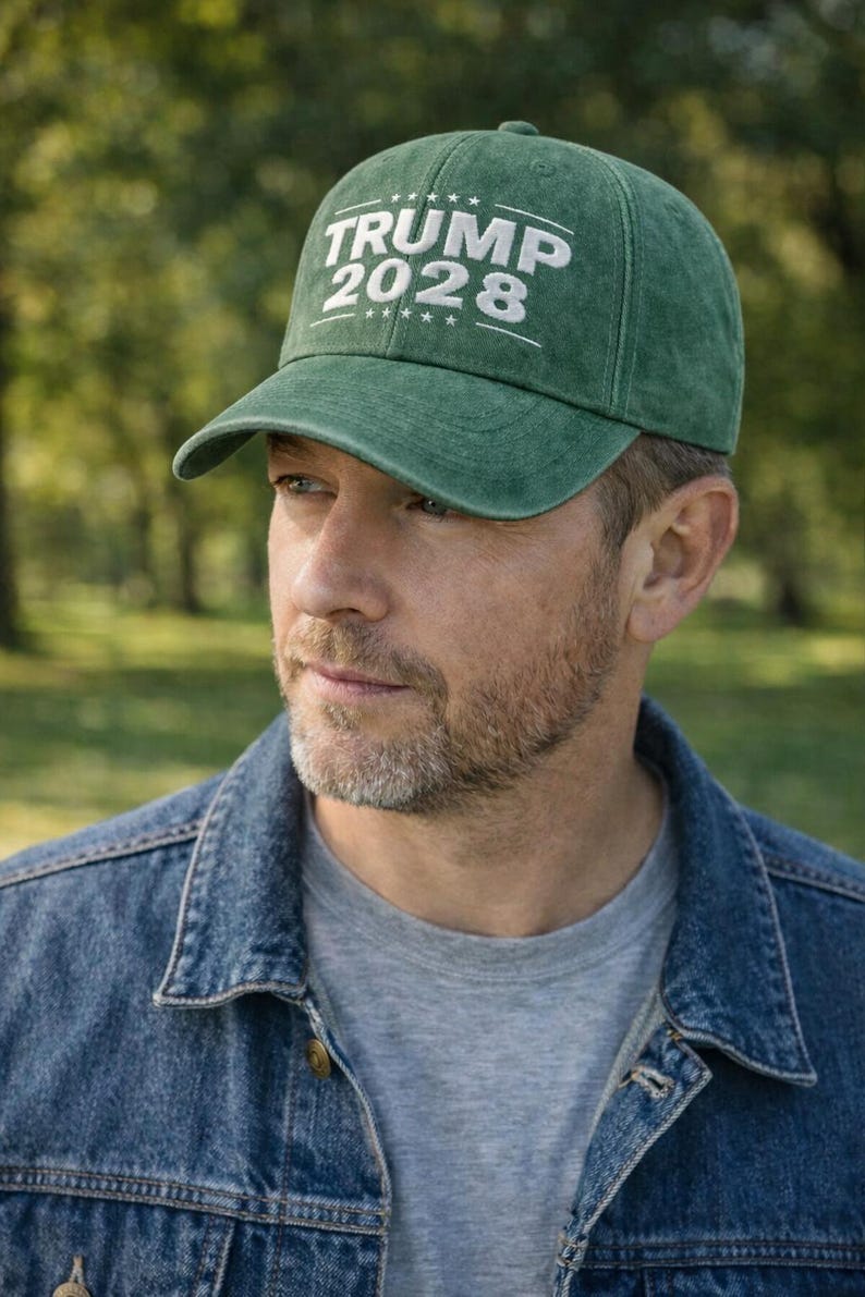 May include: Green baseball cap with the text "TRUMP 2028" in white. The cap is worn by a person wearing a denim jacket and a gray t-shirt. The background is a blurred outdoor scene.