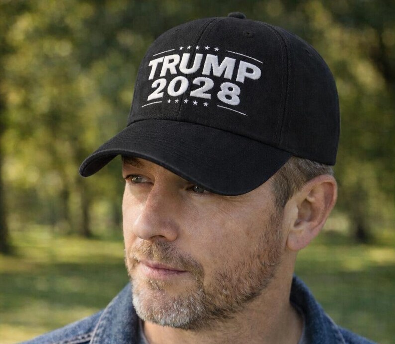 May include: Black baseball cap with the text "TRUMP 2028" in white. The cap is worn by a person wearing a denim jacket and a gray t-shirt. The background is a blurred outdoor scene with trees.