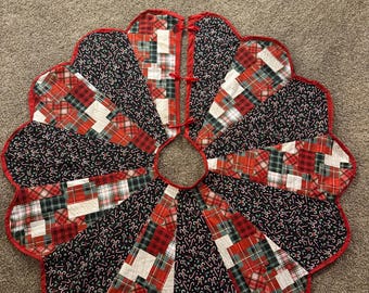May include: A circular Christmas tree skirt featuring a scalloped edge with red binding. The design consists of alternating panels of black fabric with a white candy cane print and patchwork sections in red, green, and white plaid patterns.