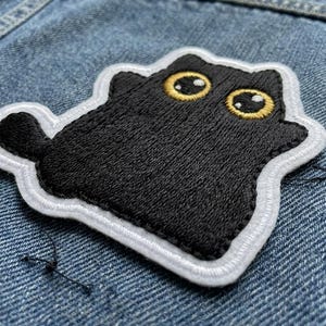 May include: Embroidered patch featuring a black cat with large yellow eyes and a white border. The patch is sewn onto a denim fabric background. The cat has a simple, cartoon-like design.