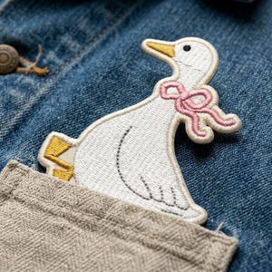 May include: Embroidered white goose patch with a pink bow, yellow beak and feet, and black eye details. The patch is sewn onto a denim jacket pocket. The jacket has a button closure.