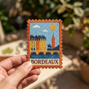 May include: A rectangular embroidered patch with a scalloped border in burnt orange. The design features a blue sky with a yellow sun, white clouds, yellow buildings, a tall tower, a dark blue bridge over water, and the text BORDEAUX in black.