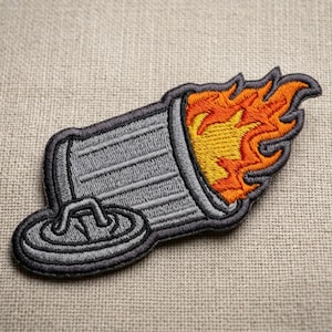 May include: Embroidered patch featuring a gray, stylized molotov cocktail with orange and yellow flames. The patch has a detailed design with a gray border and is set against a neutral fabric background.