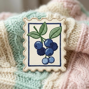 May include: A rectangular embroidered patch shaped like a postage stamp with a cream-colored background and a dark blue border. The design features a cluster of round blue blueberries with green leaves on a stem, rendered in detailed embroidery.