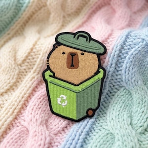 May include: An embroidered patch featuring a tan capybara with a dark brown nose and eyes peeking out from a bright green recycling bin. The bin has a matching green lid and a white recycling symbol printed on the front, all outlined in black thread.