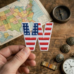 May include: An embroidered letter W patch featuring the American flag pattern with a blue field of white stars on the left side and horizontal red and white stripes across the rest of the letter, finished with a white stitched border.