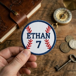 May include: A circular embroidered patch featuring a white background with red baseball-style stitching patterns. The name ETHAN is stitched in dark blue capital letters above the number 7, all enclosed within a thick dark blue circular border.