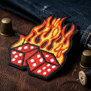 May include: Embroidered patch featuring two red dice engulfed in flames, with white dots and a black border. The flames are yellow and orange. The patch is on a brown leather surface, with denim and sewing notions in the background.