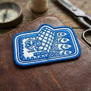 May include: A blue and white embroidered patch featuring a stylized illustration of fish and a net. The patch includes the words "SD with FRESH" in a decorative font. The patch is rectangular with a scalloped border.