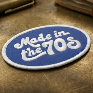 May include: Oval-shaped blue patch with white embroidered text reading "Made in the 70s." The patch has a white border and is set against a textured, golden-brown surface. A gold pen and a leather-bound book are in the background.