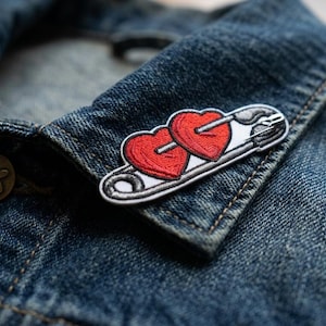 May include: Embroidered patch featuring two red hearts pierced by an arrow, set against a white and black safety pin design. The patch is affixed to a denim jacket, showcasing a casual, romantic accessory.