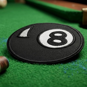 May include: A black and white embroidered patch in the shape of the number eight ball, commonly used in billiards. The patch is on a green felt surface, with a cue stick and billiard ball visible in the background.