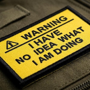May include: A yellow patch with black text that reads "WARNING I HAVE NO IDEA WHAT I AM DOING." The patch has a black border and a warning symbol. The patch is on a green fabric background.