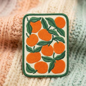 May include: A rectangular embroidered patch featuring a repeating pattern of bright orange citrus fruits with dark green leaves on a cream-colored background, finished with a dark green stitched border around the entire rectangular edge.