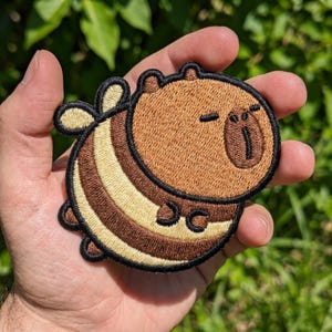 May include: An embroidered patch featuring a round, cartoon-style capybara designed with bee-like characteristics. The patch has a tan face and body with dark brown and cream-colored horizontal stripes, small wings, and a thick black stitched border.