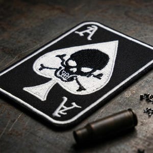 May include: Black Ace of Spades patch with a skull and crossbones design. The patch has white embroidered details and a white border. A bullet casing and metal shavings are next to the patch.