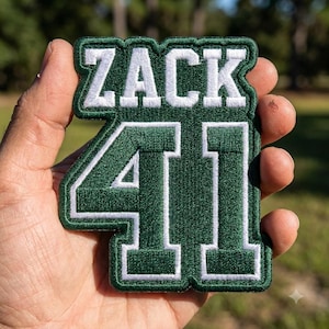 May include: A dark green embroidered patch featuring the white text ZACK above the number 41. The patch has a thick dark green border and white outlines around the letters and numbers, creating a bold, block-style design with a textured finish.