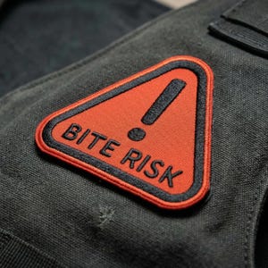 May include: A black tactical vest with a red and black embroidered patch that reads "BITE RISK". The patch features a black exclamation point inside a red triangle. The vest has black straps and a buckle.