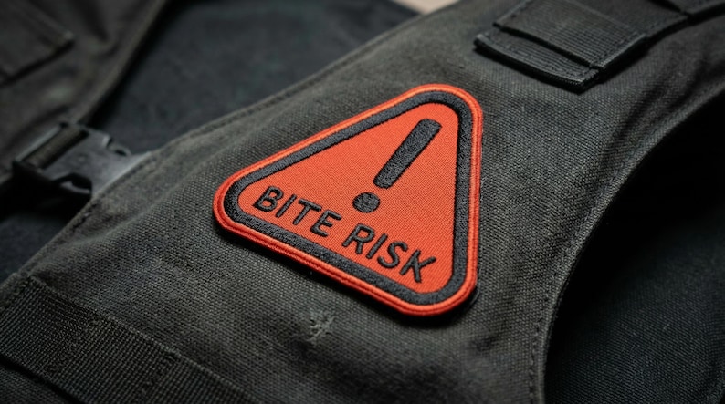 May include: A black tactical vest with a red and black embroidered patch that reads "BITE RISK". The patch features a black exclamation point inside a red triangle. The vest has black straps and a buckle.