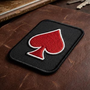 May include: Black rectangular patch featuring a red spade symbol with a white border. The patch is embroidered and set against a brown leather surface. The spade design is a classic playing card symbol.