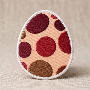 May include: Egg-shaped patch with a beige background and a pattern of maroon, red, and brown circles. The patch is outlined with a white border. This decorative patch can be sewn or ironed onto fabric.