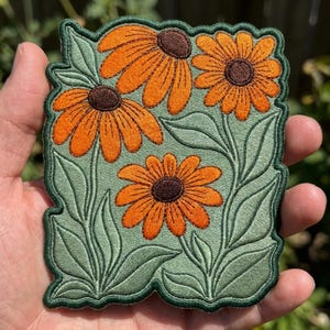 May include: An embroidered patch featuring three orange flowers with dark brown centers and detailed green leaves on a light green background. The patch has a scalloped dark green border that follows the contour of the floral design.