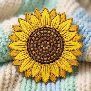 May include: A circular embroidered patch featuring a sunflower design. The center consists of a dark brown grid pattern, surrounded by two layers of vibrant yellow petals with detailed stitching that radiates outward from the center.