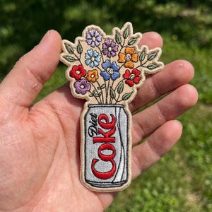 May include: An embroidered patch featuring a silver soda can with the text Diet Coke in red, topped with a colorful bouquet of embroidered flowers in shades of red, orange, blue, and purple with green leaves on a light beige fabric background.