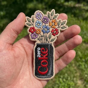 May include: An embroidered patch featuring a black soda can with the text zero Coke in red and white, topped with a colorful bouquet of embroidered flowers in shades of red, orange, blue, and purple with green leaves on a beige background.